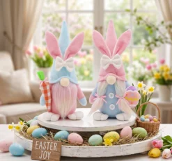 Lifestyle photograph of a pair of handcrafted Easter bunny gnomes displayed on a rustic, decorated wooden tray. The gnomes feature long ears and bowties; the left gnome holds a checkered carrot, the right holds a patterned Easter egg. The tray is filled with straw, colorful assorted Easter eggs, spring flowers, a small basket, and a wooden sign that reads "EASTER JOY". Background is a cozy, sunlit living room with a garden view.